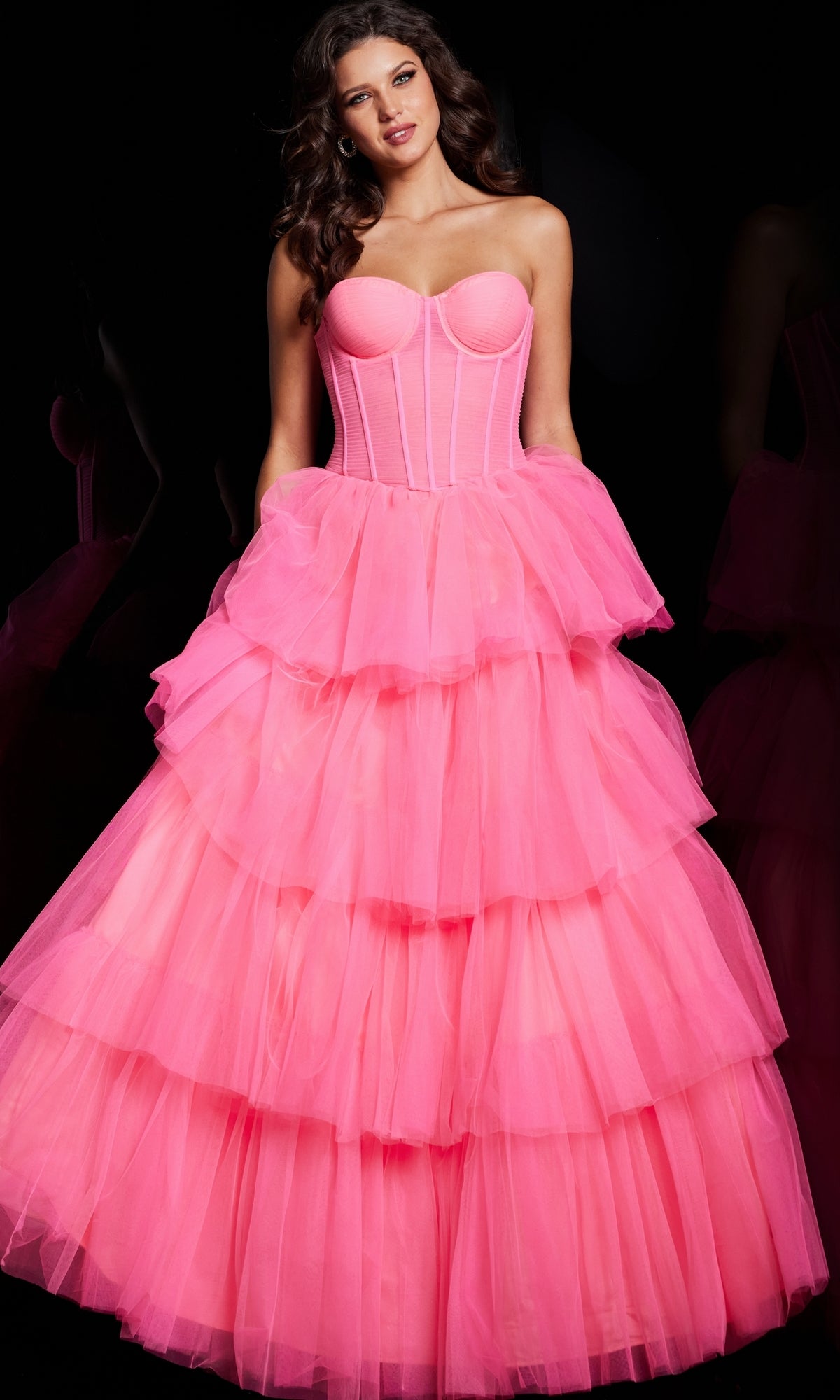 A woman stands against a black background wearing the Jovani 37062 Strapless Long Ruffle Prom Ball Gown in hot pink with a corset-style bodice and tiered tulle skirt.